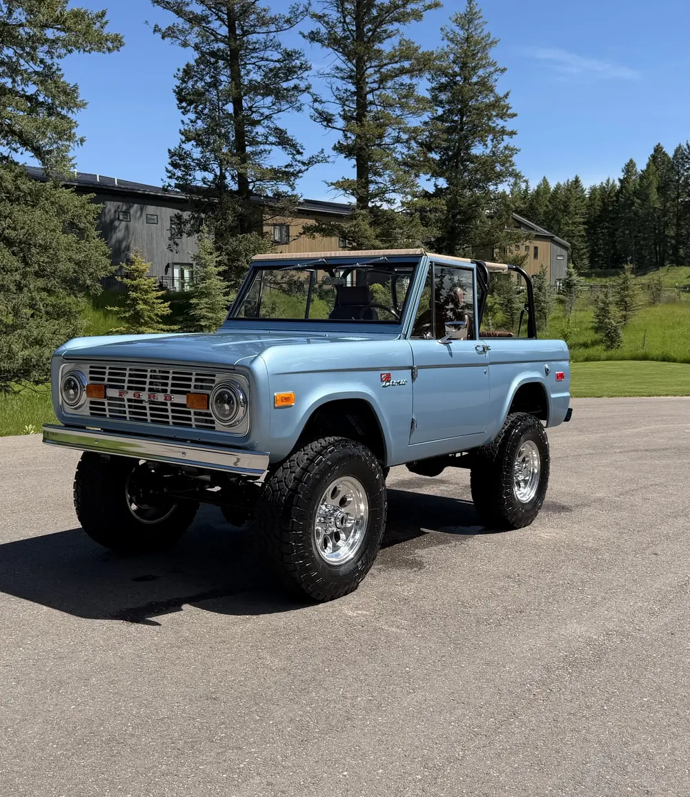 Light blue classic Ford Bronco with the top off, parked under tall pines after a detail by Stumptown Detailing